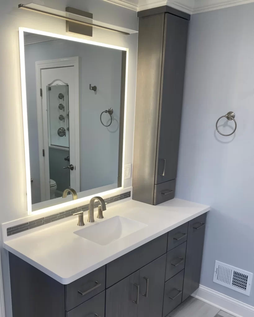 Custom gray bathroom vanity in a Berwyn, PA primary bathroom remodel with an illuminated mirror, tall storage cabinet and brushed metal fixtures.
