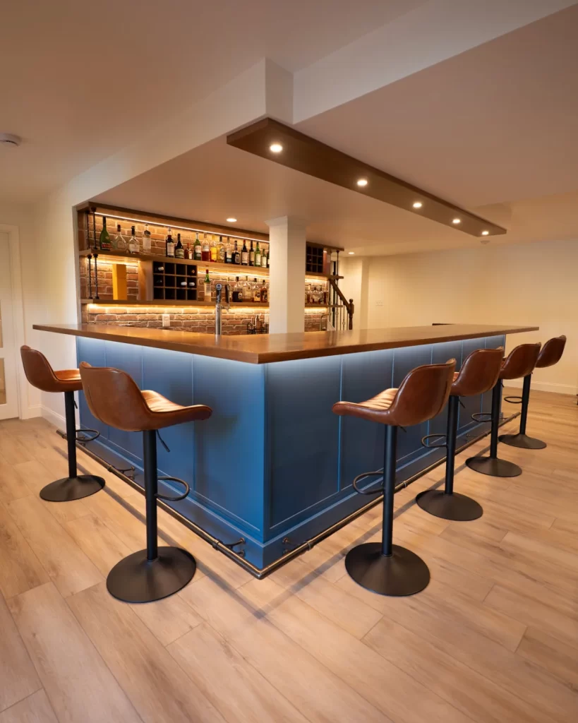 Custom basement wet bar in West Chester, PA with blue cabinetry, wood bartop, bar seating, illuminated shelving, and built-in footrest.