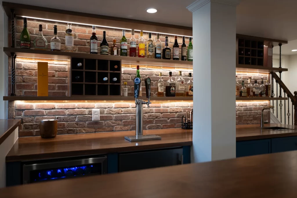 Custom basement bar in West Chester, PA with wood shelving, brick backsplash, integrated lighting, cabinetry, and beverage storage.