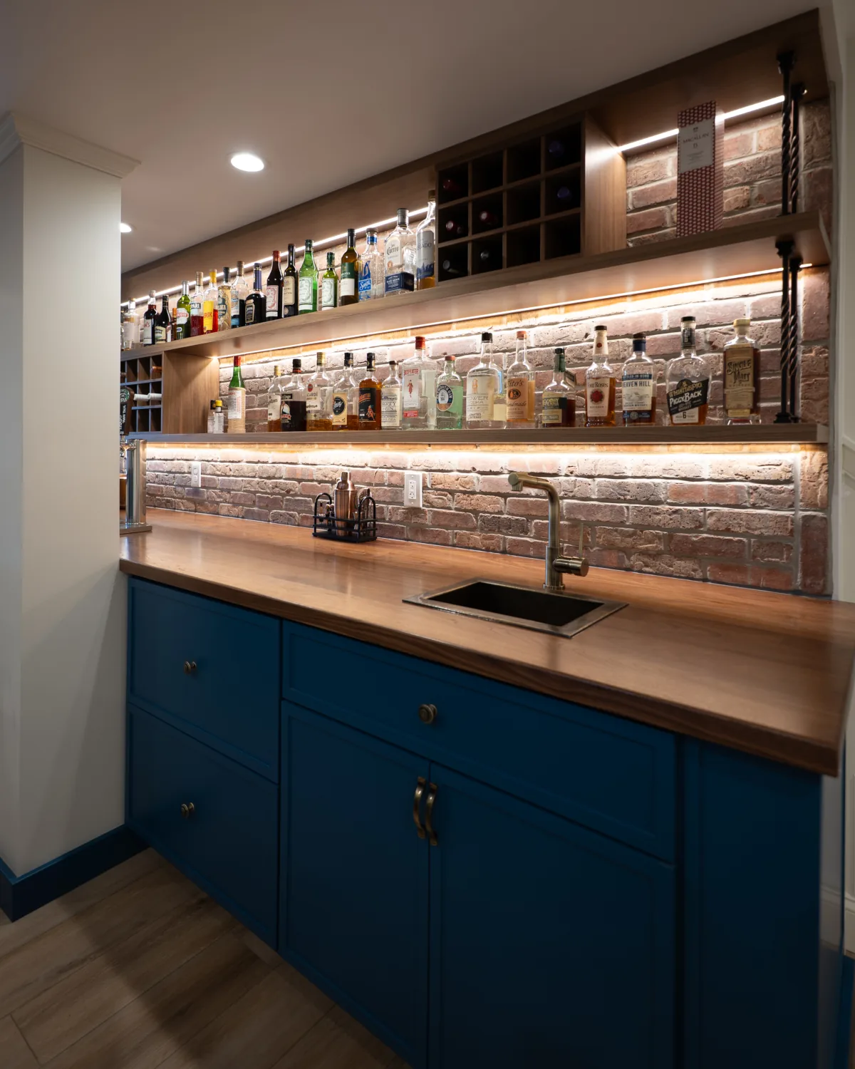 Custom wet bar in a West Chester, PA basement remodel with blue cabinetry, wood countertop, brick backsplash, sink, and illuminated wood shelving.