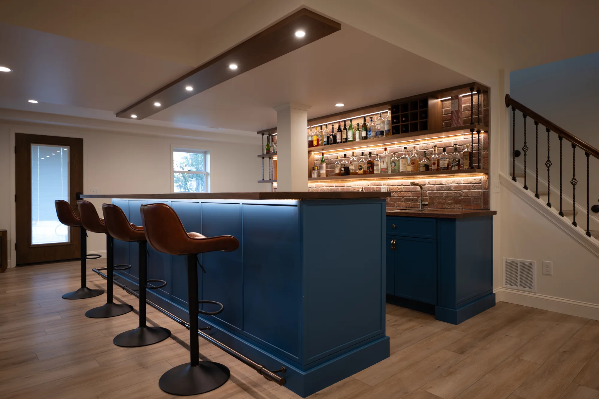 Finished basement remodel in West Chester, PA with custom wet bar, built-in shelving, bar seating, wood flooring, and integrated lighting.