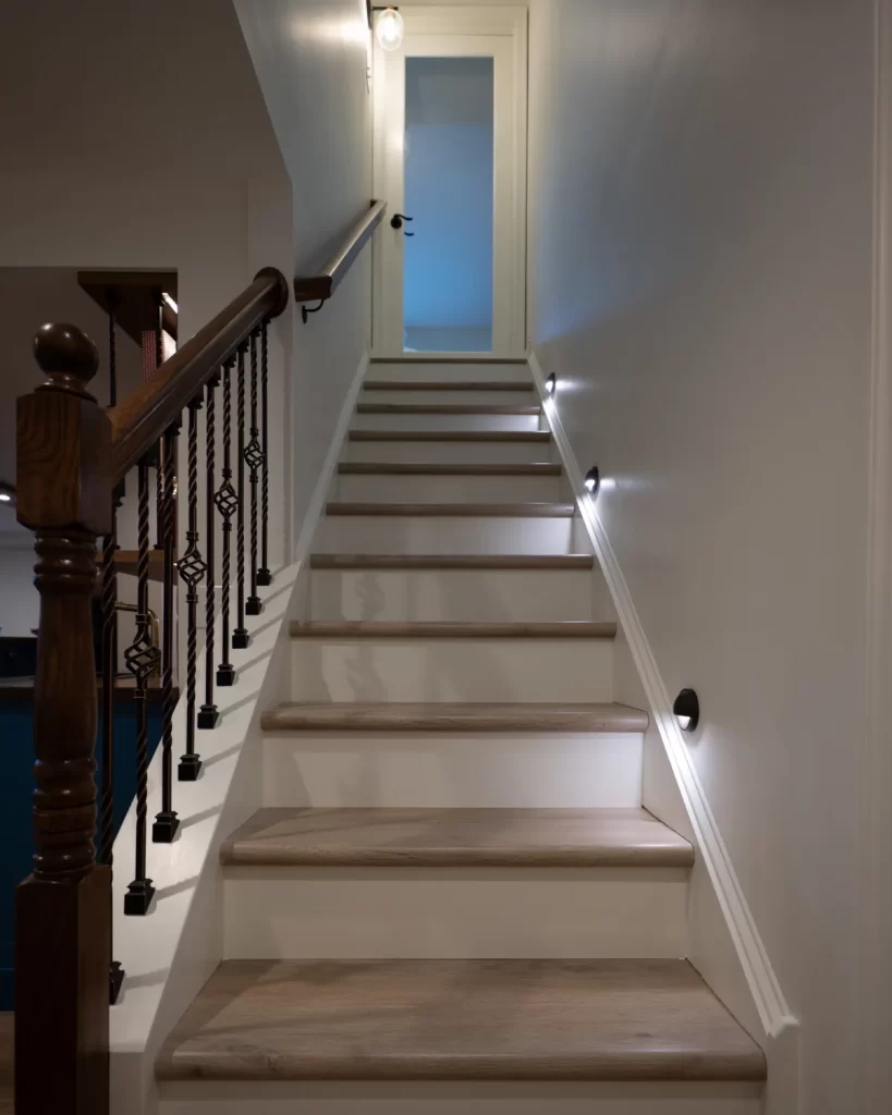 Finished basement staircase in West Chester, PA with updated stair treads, wood railing, iron balusters, wall lighting, and basement door.
